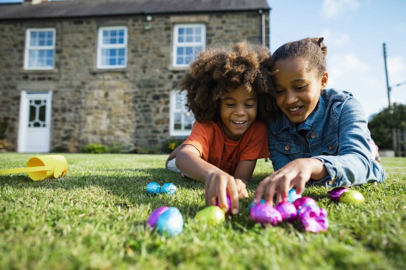 A front-view shot of a young boy and his older sister, they are lying down on their front smiling while holding their Easter eggs in front of their holiday cottage.