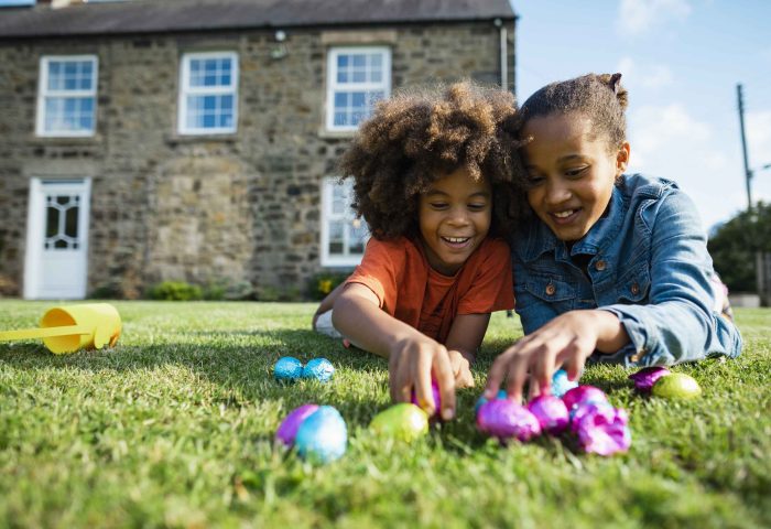 A front-view shot of a young boy and his older sister, they are lying down on their front smiling while holding their Easter eggs in front of their holiday cottage.